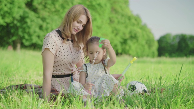 快乐的年轻妈妈帮助可爱的小女孩吹肥皂泡。快乐的白人女儿花周末与母亲在阳光灿烂的绿色夏季草地上的肖像。快乐,儿童,生活方式视频素材