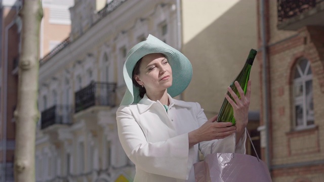 中景肖像快乐优雅的女人在户外欣赏一瓶白葡萄酒。戴着帽子微笑的白人女士手里拿着酒站在城市街道上。休闲的生活方式,财富。视频素材