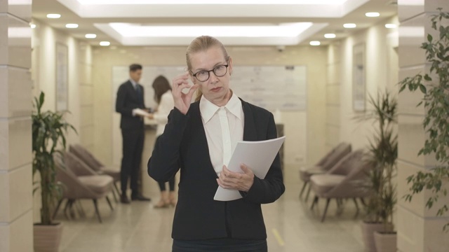 一幅严肃的中年女商人戴着眼镜在大厅里拿着文件摆姿势的肖像。自信的职业白人女性站在商业中心看着镜头。电影4k ProRes总部。视频素材