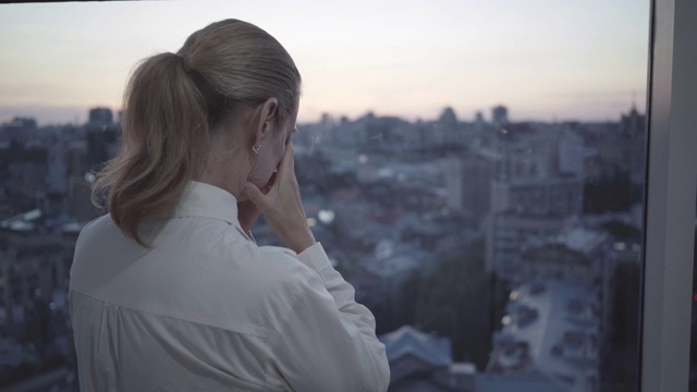 沮丧的中年女商人站在窗前看着夜晚的城市。在黄昏时分站在阳台上思考的紧张的白人妇女的背影。超负荷工作的概念。视频素材