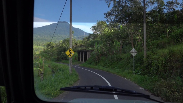 视频从前面的汽车行驶在巴厘岛的道路弯曲的道路，印度尼西亚在阳光明媚的日子里，倒下的树木排列的道路和山景。视频素材