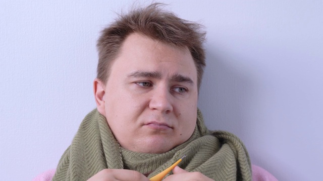 一个悲伤的人戴着暖和的围巾，嘴里叼着温度计，测量体温，然后举手认输。一名患有流感和发烧的年轻人被隔离在家中。视频素材画面2