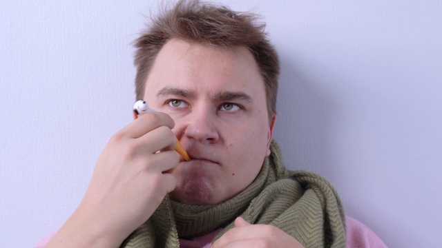 一个悲伤的人戴着暖和的围巾，嘴里叼着温度计，测量体温，然后举手认输。一名患有流感和发烧的年轻人被隔离在家中。视频素材画面4