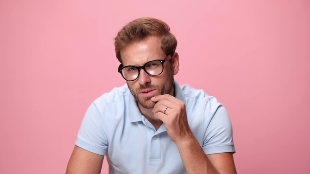 性感的年轻休闲男人在蓝色polo衫触摸性感的下巴和思考，固定眼镜和看向一边，微笑和在粉红色的背景调情视频素材