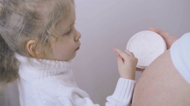 金发女孩从罐子里把乳液涂在怀孕母亲的肚子上视频素材