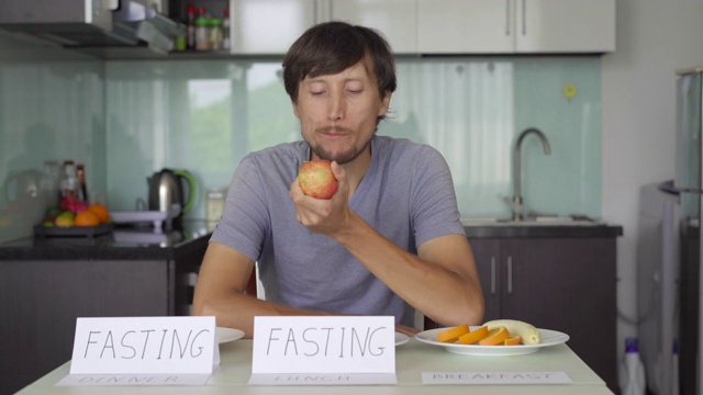 一个悲伤的年轻人把禁食的标志放在空盘子上，标志午餐和晚餐，然后从早餐盘子里吃了一个苹果。间隔禁食的概念。不吃饭视频素材画面2