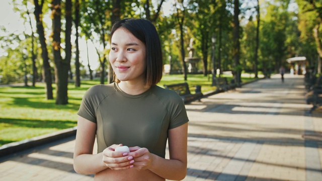 户外肖像的年轻亚洲女士把无线耳机在公园和微笑，跟踪拍摄视频素材