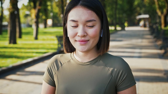 户外拍摄的年轻亚洲女性，在耳机里听音乐，在公园里对着镜头微笑，跟踪拍摄视频素材