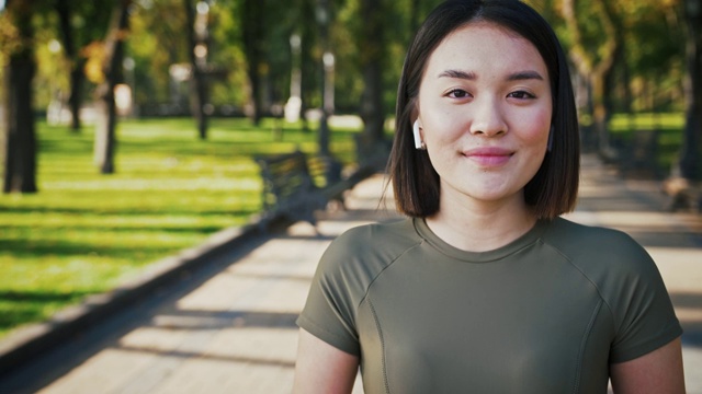 可爱的亚洲女人戴着无线耳机在城市公园对着摄像头微笑，跟踪拍摄视频素材