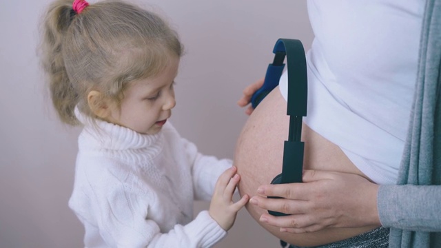 有趣的女孩和怀孕的妈妈把耳机放在肚子上视频素材