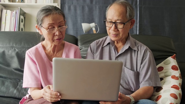 亚洲老夫妇，爷爷和奶奶在家里客厅的沙发上用笔记本电脑上网。老年人学习在生活方式活动中使用新科技。视频素材