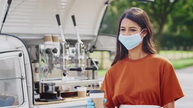 在卫生大流行期间，戴口罩的妇女在接待女性顾客的面包车上经营独立移动咖啡店-慢镜头视频素材