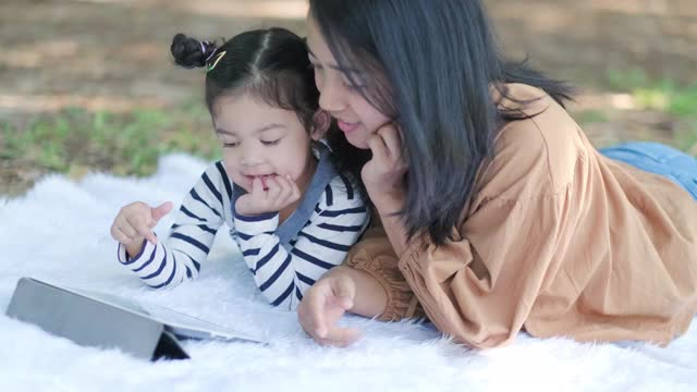 亚洲的母亲躺在地板或地面上和她的女儿在花园里玩平板设备。妈妈教她如何使用。与孩子交谈。现代电子技术与家庭观念。感觉快乐视频素材