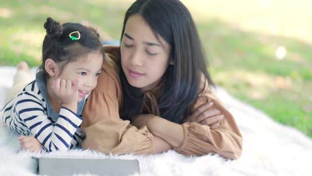 亚洲的母亲躺在地板或地面上和她的女儿在花园里玩平板设备。妈妈教她如何使用。与孩子交谈。现代电子技术与家庭观念。感觉快乐视频素材