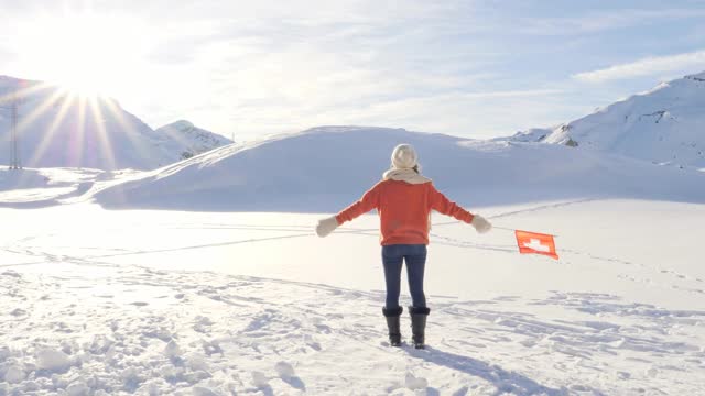 一个年轻的女人站在高高的雪山周围，在空中举着瑞士国旗。女人张开双臂举着瑞士国旗。慢动作视频素材