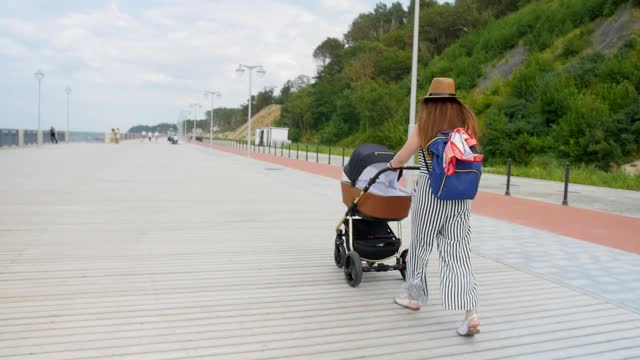 快乐的年轻妈妈推着婴儿车在散步。家庭，母亲和人的概念-快乐的母亲与婴儿睡在婴儿车散步在散步的夏天。全景。视频素材