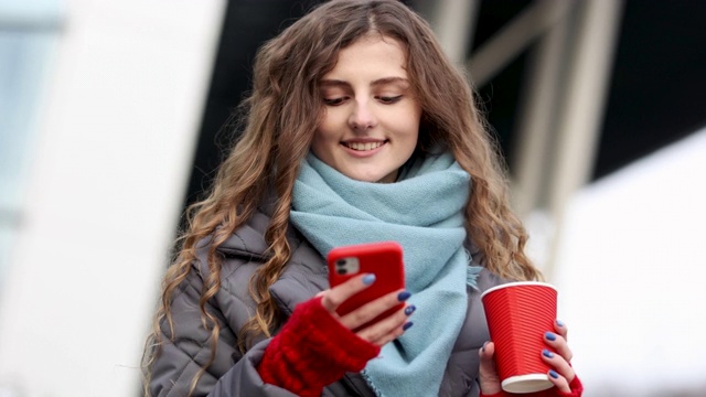 肖像幸福美丽的卷发女人用咖啡杯外卖用手机发短信。女人在冬天的街道上四处张望。4 k慢动作视频素材