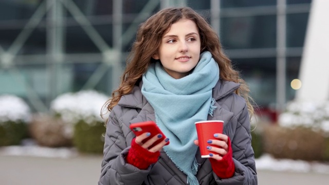 肖像幸福美丽的卷发女人用咖啡杯外卖用手机发短信。在街道上四处走走。4 k慢动作视频素材