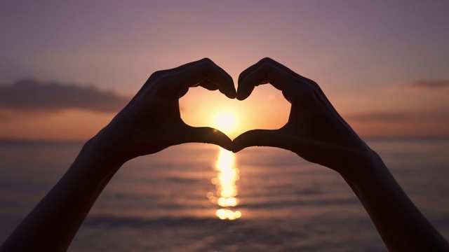 在海上美丽的日落上，女人用手做出心形。视频购买
