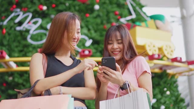 两位快乐的亚洲年轻女性在百货商店的户外购物在销售季节在年底。女孩们拿着购物袋，快乐地走着，微笑着一起买东西视频素材