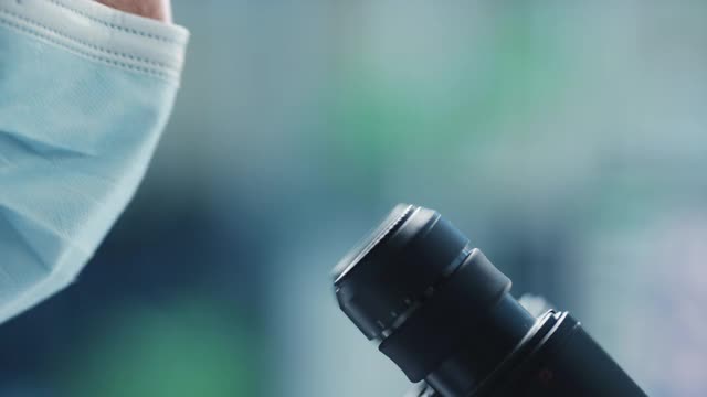 一个英俊的男性科学家戴着医用口罩和在显微镜下看的宏近镜头。在现代实验室用技术设备研究分子样本的微生物学家。视频素材