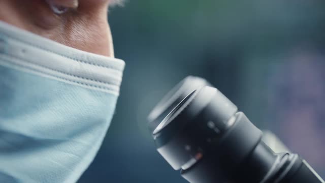 一位男性科学家戴着医用口罩并在显微镜下观察的超微距特写镜头。在现代实验室用技术设备研究分子样本的微生物学家。视频素材