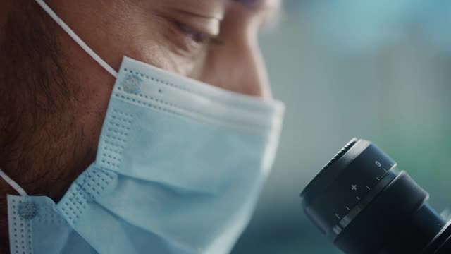 一个英俊的男性科学家戴着外科口罩和看进显微镜的宏观近镜头。在现代实验室用技术设备研究分子样本的微生物学家。视频素材
