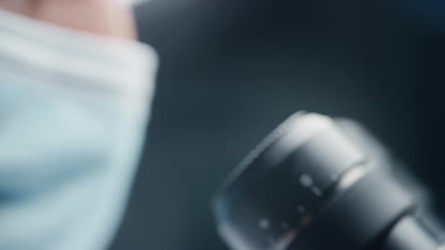 一位男性科学家戴着医用口罩并在显微镜下观察的超微距特写镜头。在现代实验室用技术设备研究分子样本的微生物学家。视频素材