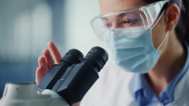 微距镜头的一个女科学家在面罩和护目镜看显微镜。在现代实验室中使用技术设备研究分子样本的女性微生物学家。视频素材