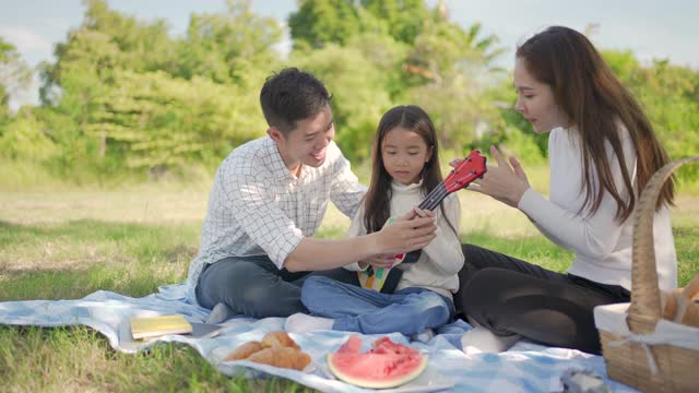 快乐的亚洲家庭和小女孩玩尤克里里，并享受了我们一起在野餐布在绿色花园。一家人在大自然中享受阳光明媚的秋日。视频素材