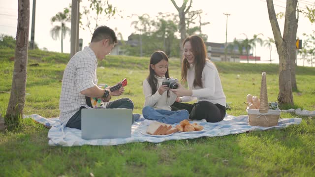 幸福的家庭亚洲和小女孩玩尤克里里，而父亲拍照享受我们一起在野餐布在绿色花园。一家人享受阳光明媚的秋日。视频素材