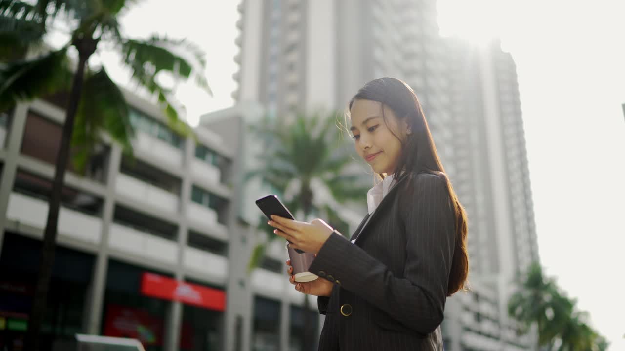 视频缓慢。亚洲女人，漂亮的女商人，白人，穿着黑色西装，在用智能手机聊天。在城市街道的一边视频素材