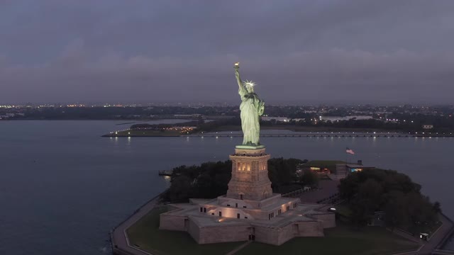 美丽的自由女神像在日出或黄昏与美国国旗挥舞的背景，空中滑左多莉视频素材