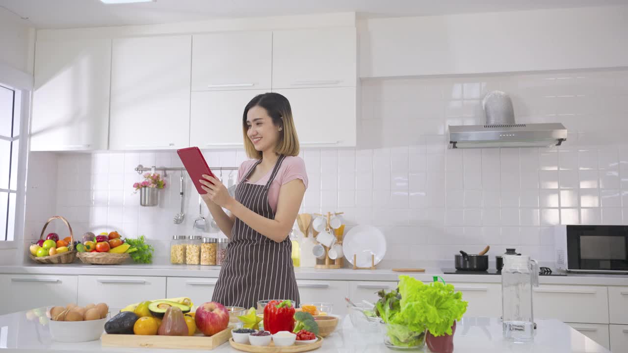 亚洲妇女用手指在平板电脑屏幕上滑动准备烹饪原料，在家庭厨房的网站上跟随烹饪在线视频剪辑。厨房里有健康蔬菜的女人。视频素材