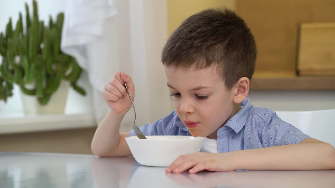 饥肠辘辘的小男孩在早晨吃着香甜的玉米片和牛奶。一个小男孩独自坐在餐桌旁。视频素材