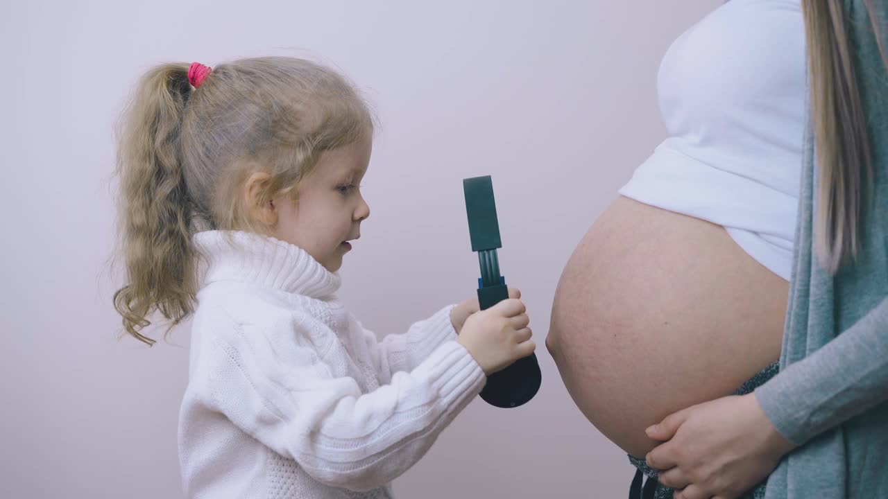 可爱的女孩把耳机戴在怀孕妈妈的肚子里视频素材