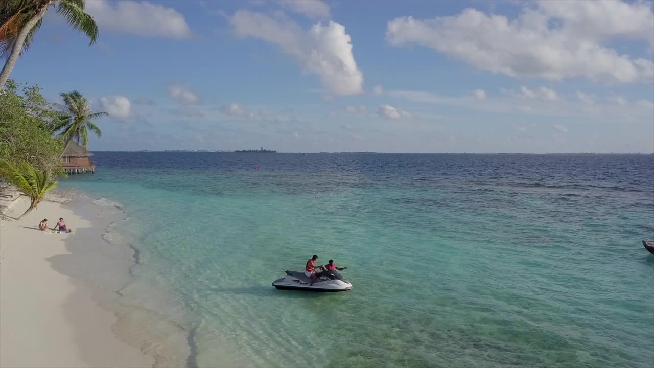 阳光明媚的一天，马尔代夫马累环礁，游客在海滩上玩水上摩托艇，无人机在天空中靠近人群视频素材