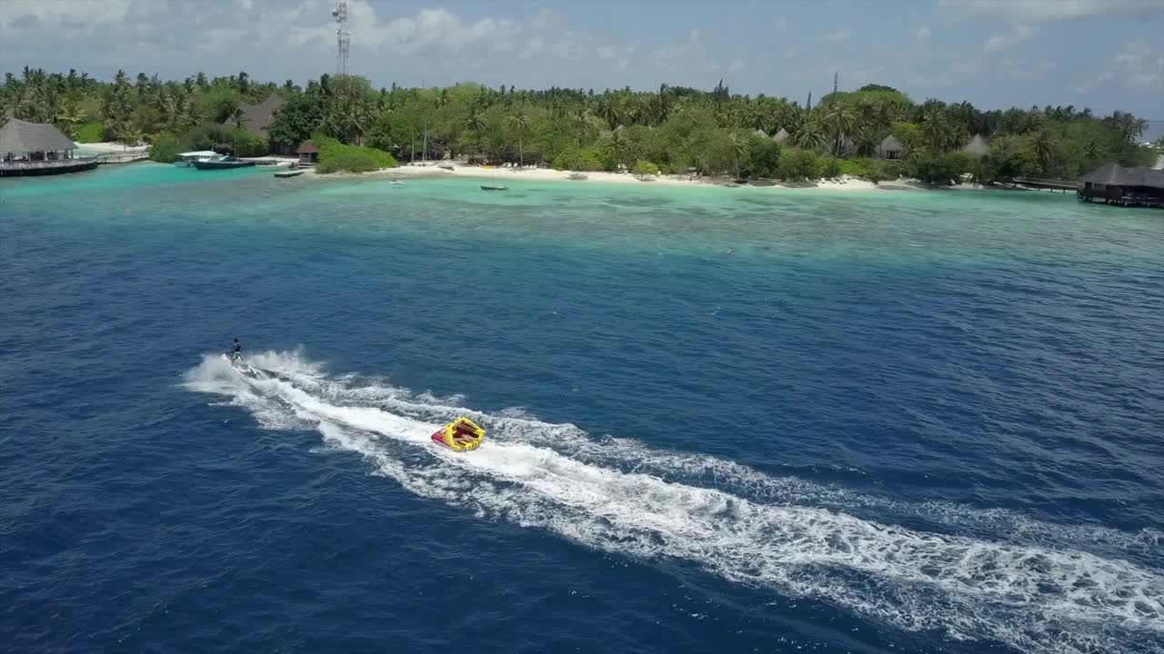 空中摇摄水上摩托艇拉有趣的管子与游客在海上，无人机飞过附近的海滩对天空在阳光明媚的一天-南马累环礁，马尔代夫视频素材