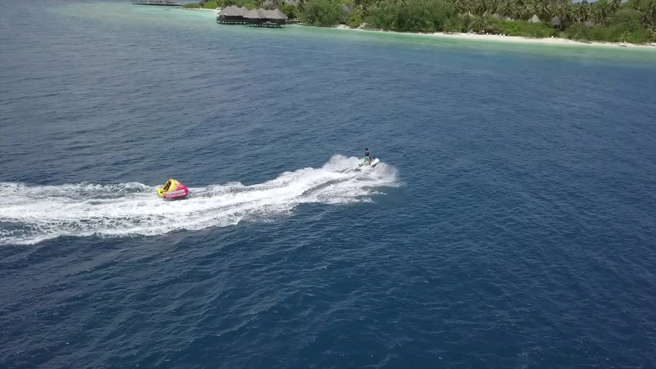 空中摇摄喷气式滑雪拉有趣的管子在蓝色的海，无人机飞越游客享受水上运动在阳光明媚的一天-南马累环礁，马尔代夫视频素材