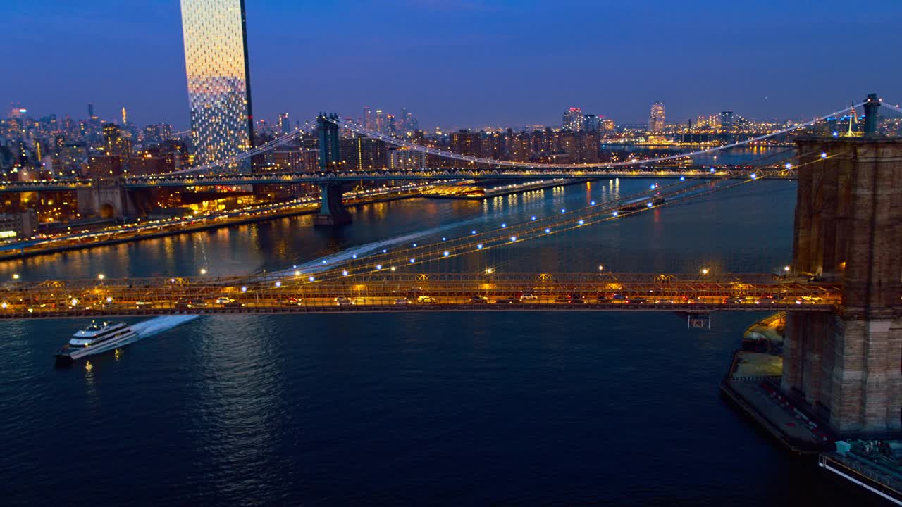 晚上的车流中，水轮从布鲁克林大桥(Brooklyn Bridge)下驶过，车水马龙，背景是曼哈顿中城(Midtown Manhattan)的天际线，在夜色中灯火通明。无人机视频与向后平移摄像机运动。视频素材