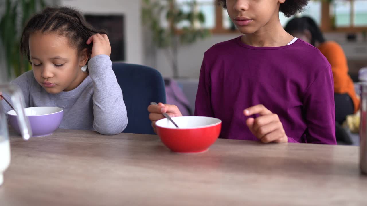 两个可爱的非洲女孩吃麦片早餐，而他们的母亲正在客厅放松视频素材