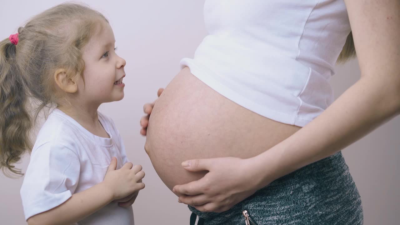 快乐的女孩抱着怀孕的妈妈的肚子站在墙上玩视频素材