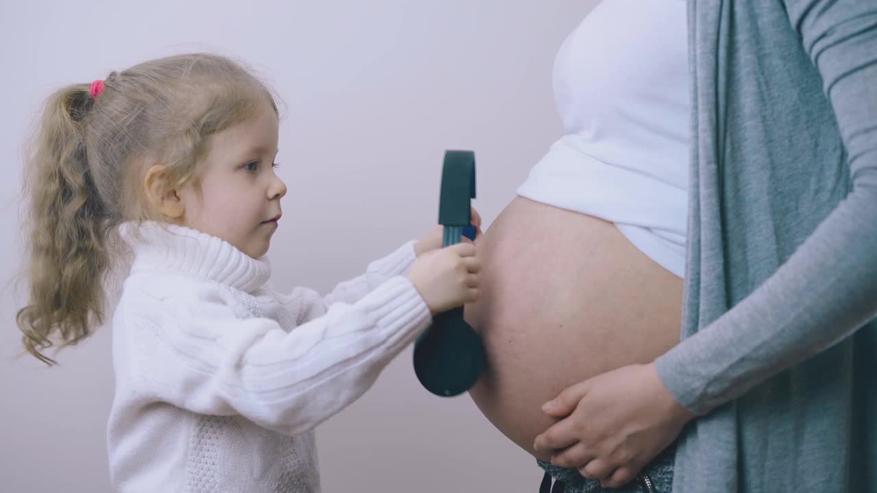 有趣的女孩戴着耳机把头放在怀孕妈妈的肚子上视频素材