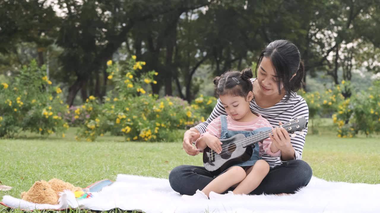 亚洲女人玩尤克里里与她可爱的女儿在公园野餐。视频素材