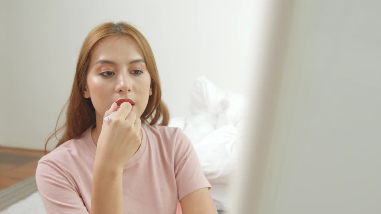 近距离肖像迷人的亚洲女孩胭脂她的嘴唇。她拿着粉红色的口红。她的嘴轻轻张开，看着镜子。视频素材