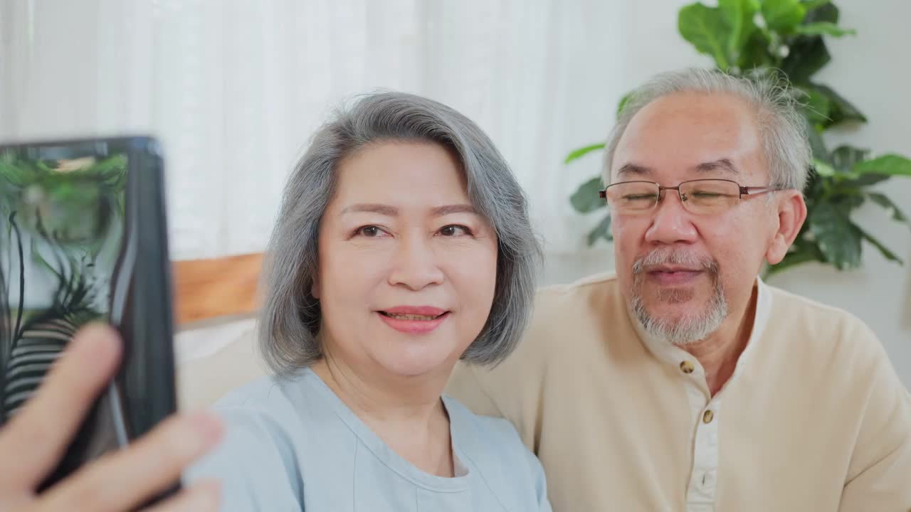 亚洲老年夫妇的特写，老年妇女和老年男子使用手机视频通话与儿子，女儿和在家里与孙辈交谈。祖父母对通过互联网交流感到高兴视频素材