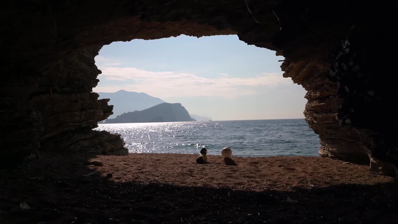 在阳光明媚的日子里，在宁静的海水附近的沙滩上，达克斯猎狗罗宾逊和球朋友在洞穴入口处的剪影视频素材