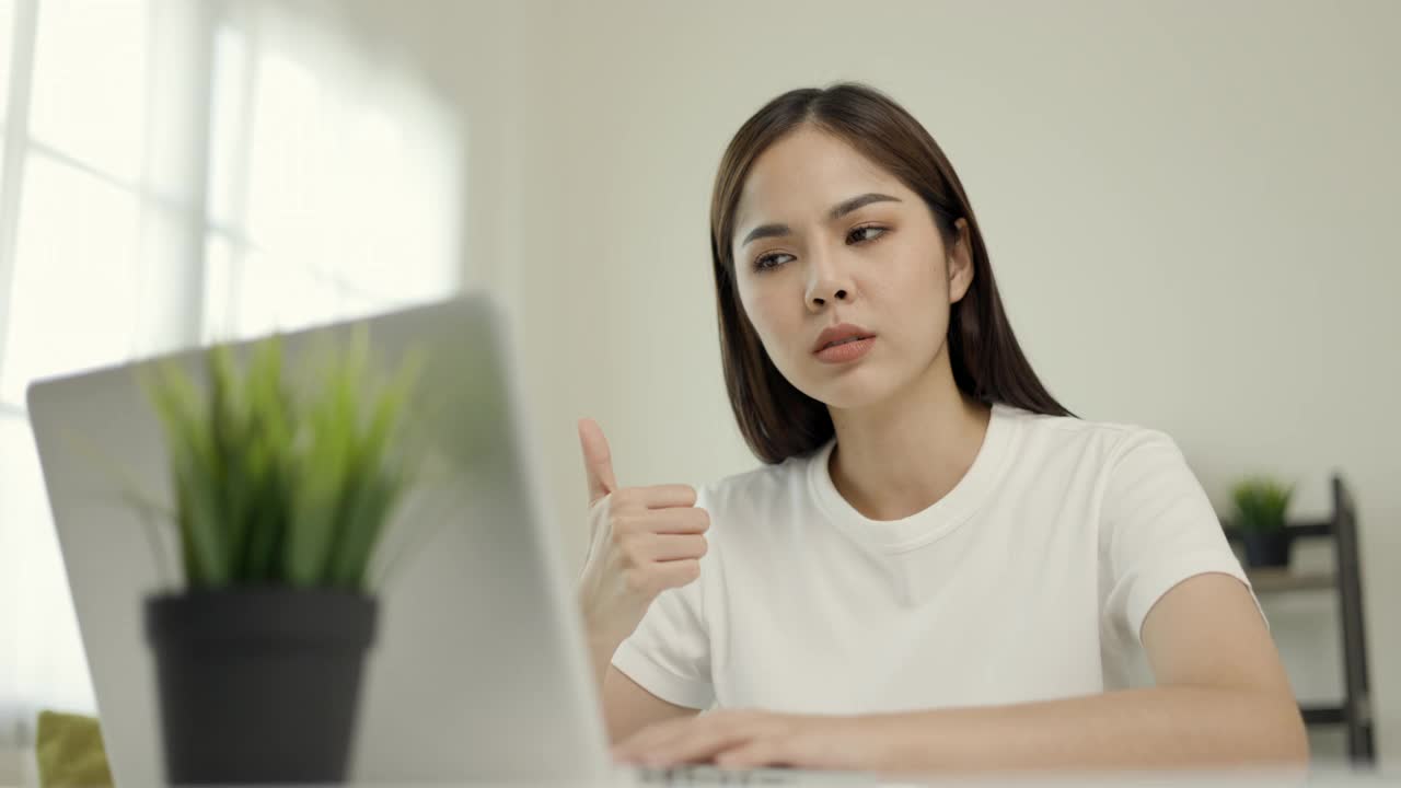 年轻的亚洲女子坐在家里的客厅里用笔记本电脑聊天视频在线会议。商务女性看屏幕在社交媒体上的实时蒸汽会议。工作，在家学习。视频素材