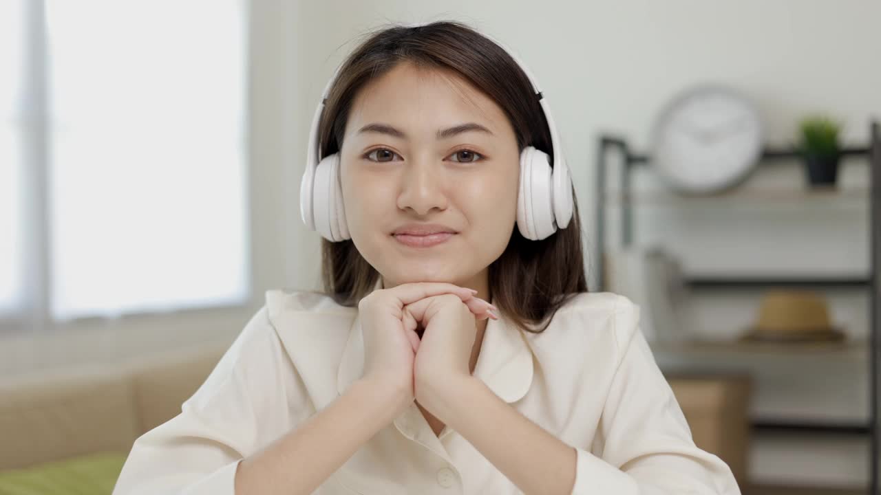 年轻的亚洲女子戴着耳机看着摄像头坐在家里的客厅里在线视频会议。商业女性看屏幕会议在社会媒体直播蒸汽。工作，在家学习。视频素材