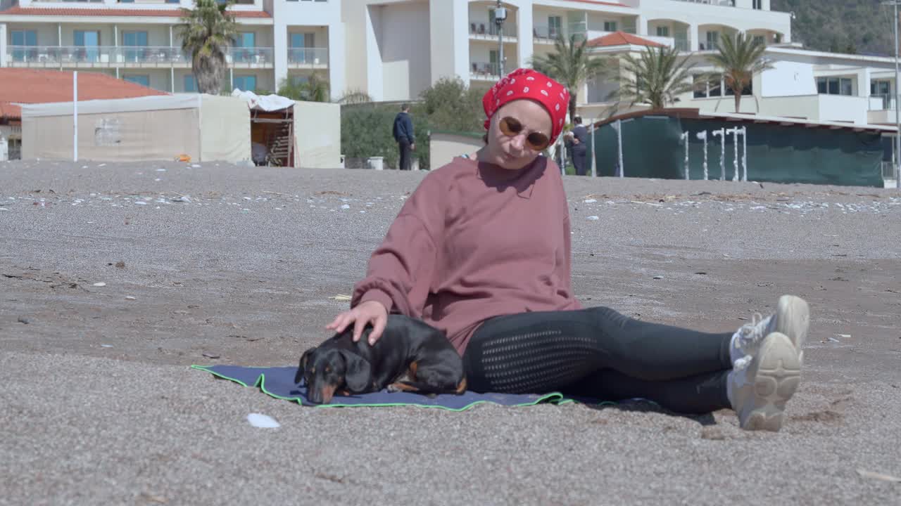 阳光明媚的春日，戴着太阳镜的漂亮女人抚摸着小腊肠犬在度假酒店附近的沙滩上消磨时光视频素材